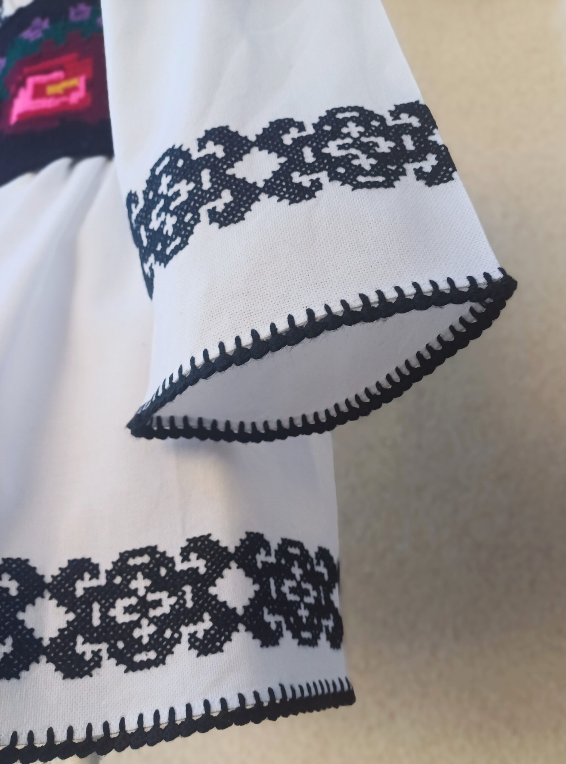 „Costum tradițional moldovenesc brodat 'Nicu' din zona Sucevei, confecționat din pânză albă de bumbac, cu broderie geometrică neagră și brâu țesut cu motive florale colorate.”